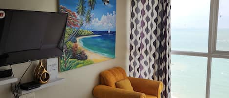 A room with a large window, a painting of a beach, a yellow sofa, and a television on a shelf.