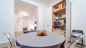 Un coin repas avec une table ronde, des chaises transparentes, un panier de fruits et une bibliothèque contenant divers objets.