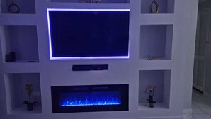 A modern living room with a flat-screen TV mounted on a white wall, surrounded by shelves. Below the TV, there's a built-in fireplace with a blue flame.