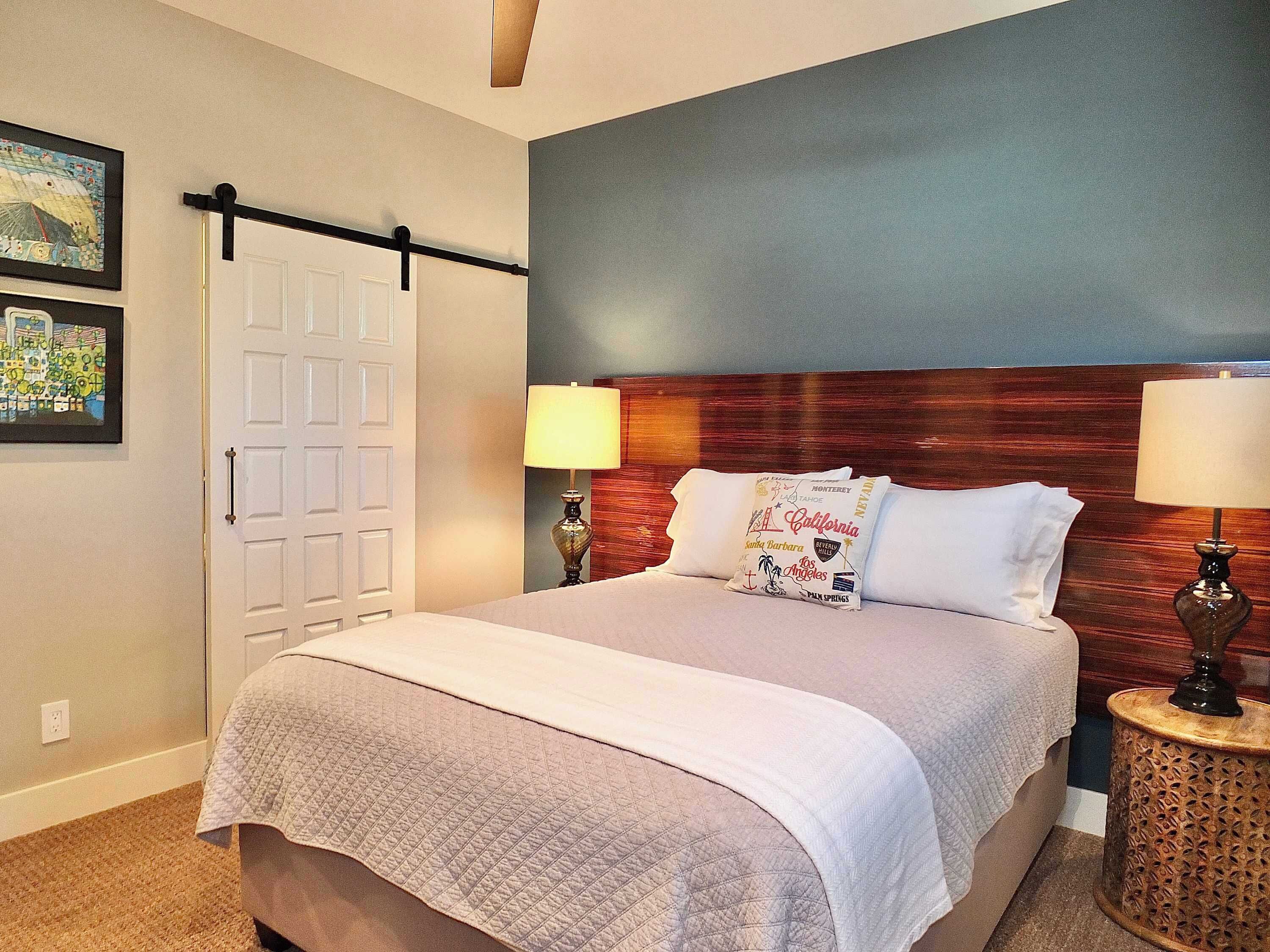 Un lit bien fait, avec une couverture matelassée, deux lampes de chevet et une tête de lit ornée d’un coussin sur le thème de la Californie.