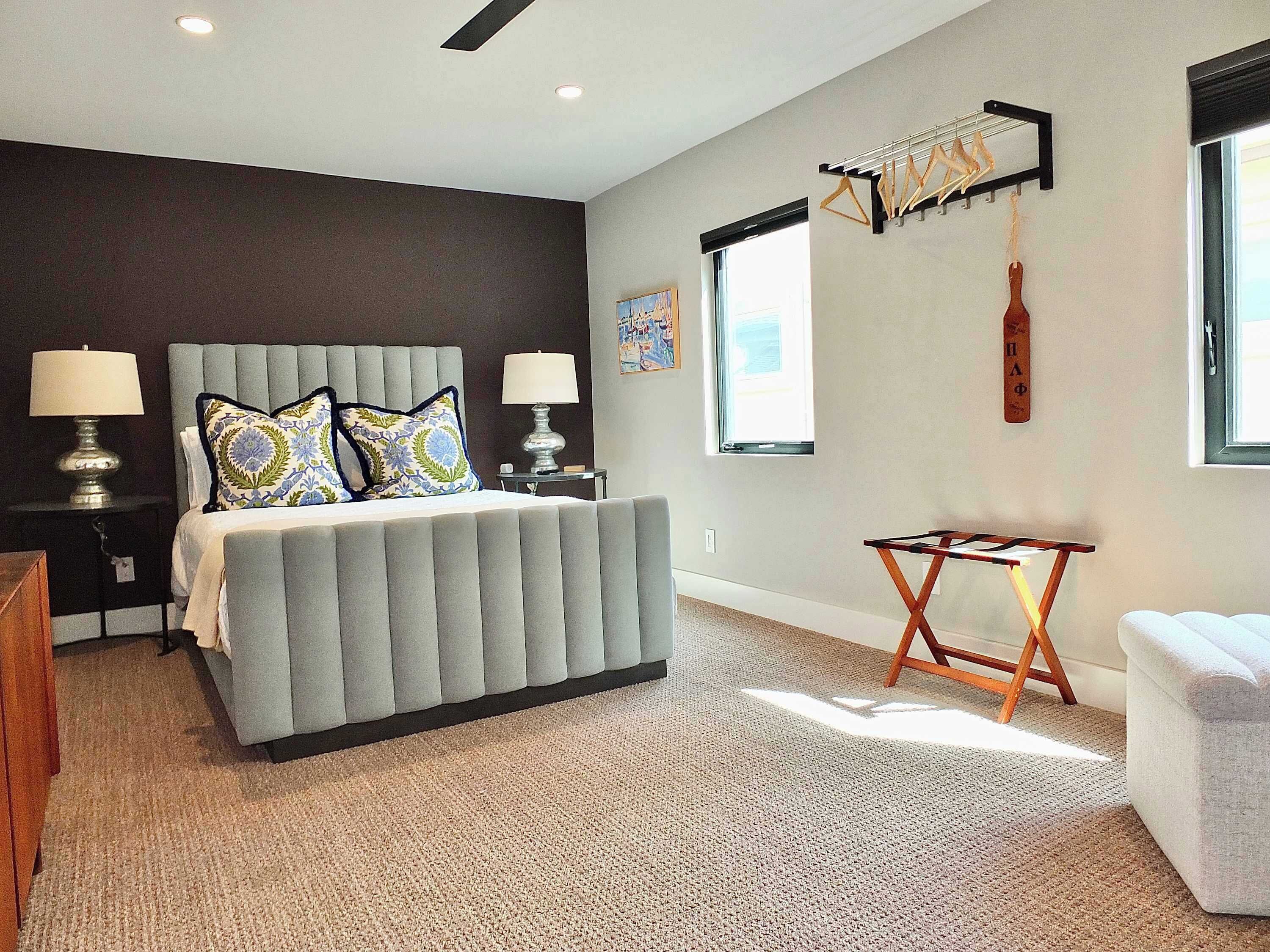 A modern bedroom with a large bed, two bedside tables with lamps, a small wooden side table, a window with a view, and a wall-mounted drying rack.