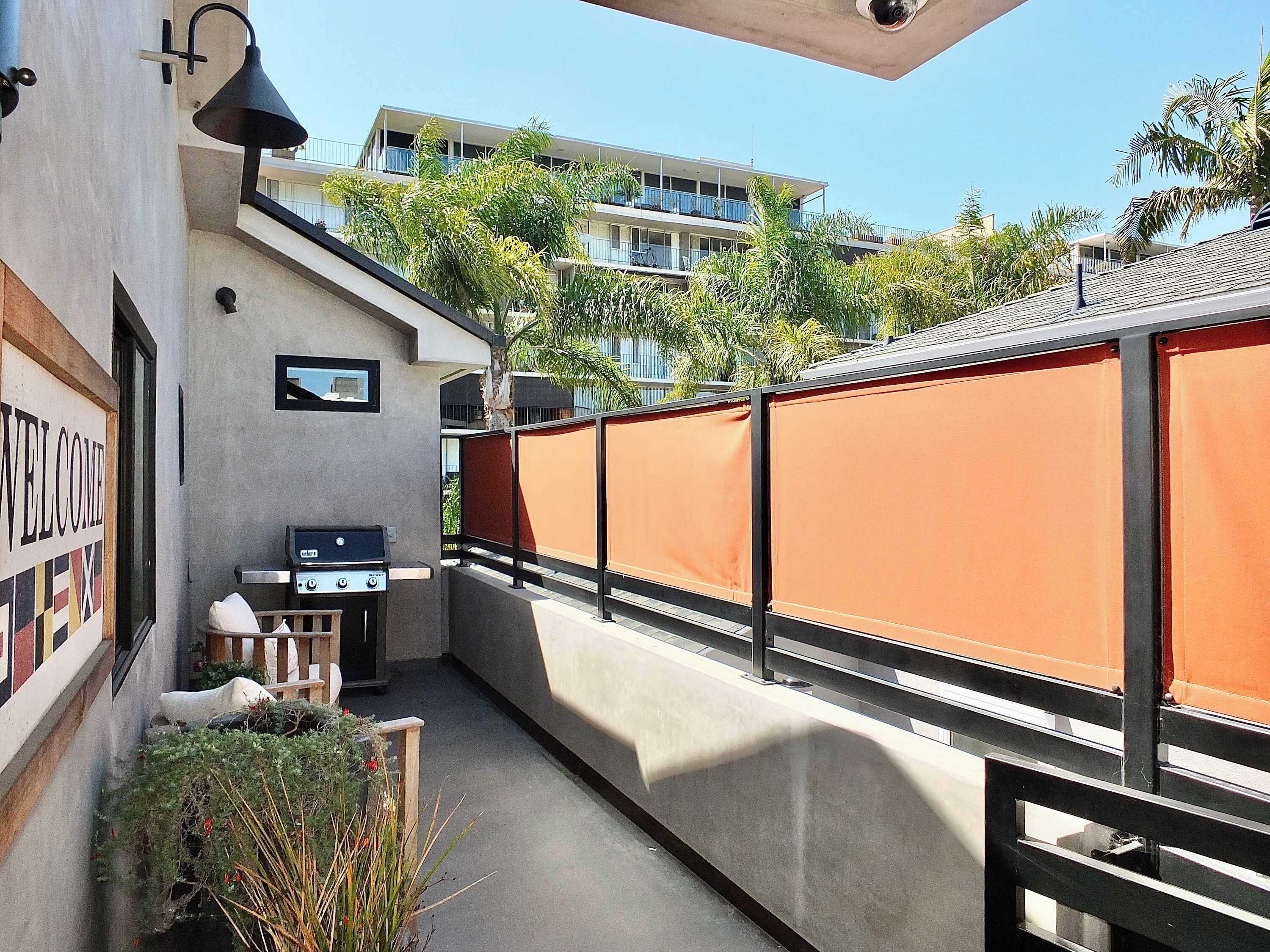 A balcony with a barbecue grill, a welcome sign, and a view of a multi-story building with balconies.