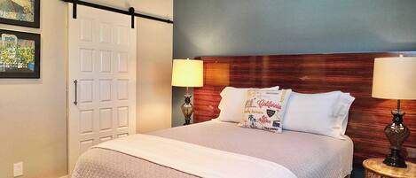Una cama bien tendida con cubrecolcha acolchado, dos lámparas de noche y un cabecero con una almohada alusiva a California.