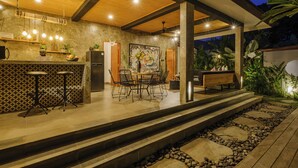 An outdoor patio area with a dining table and chairs, a bar counter with stools, and a stone pathway leading to the entrance. - Villa Gaya Ubud (Ubud)