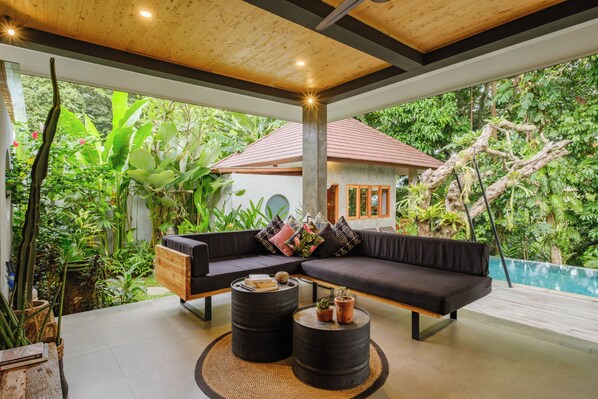 A modern outdoor living area with a wooden ceiling, a black sofa with cushions, two round tables, and a view of a house and lush greenery.