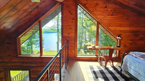 Cabana com grandes janelas que oferecem vista para um lago e uma floresta. Há uma cama, uma escrivaninha e uma cadeira no quarto.