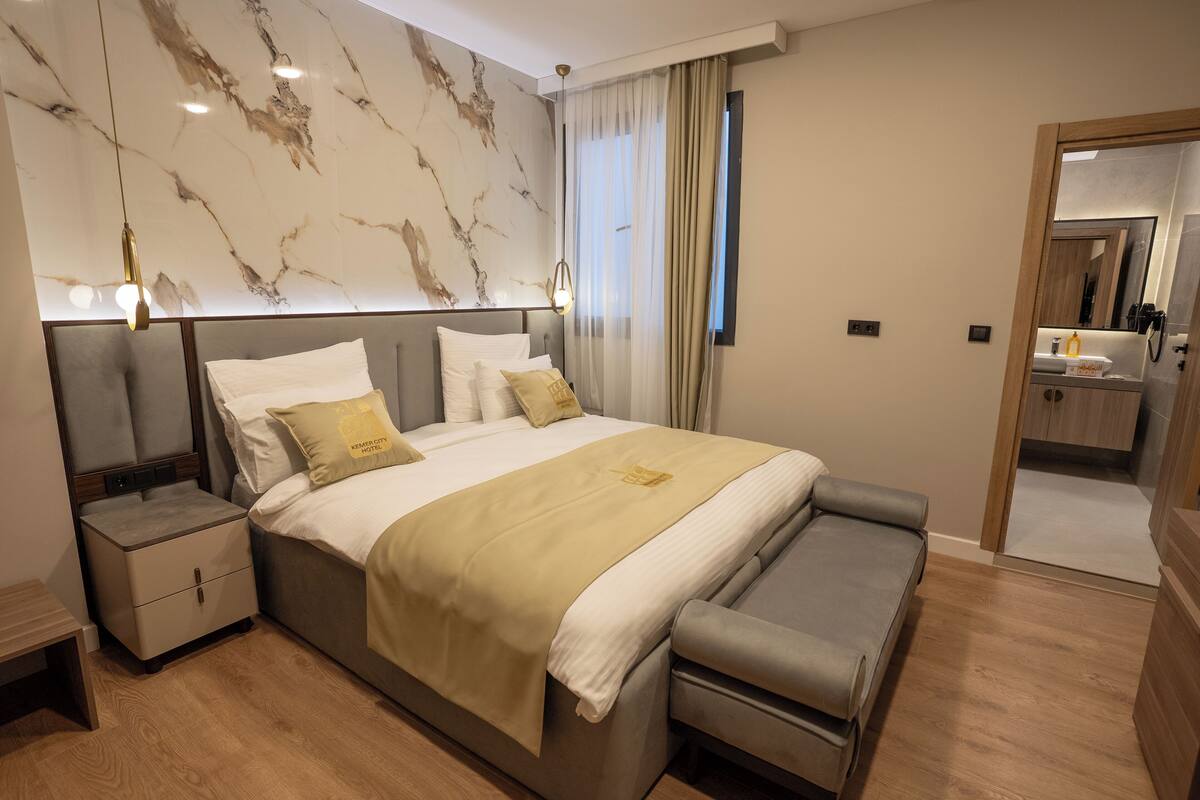A modern bedroom with a large bed, bedside tables, a bench, a marble wall, and a bathroom visible through an open door.