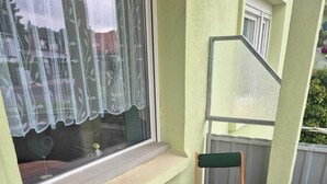 Un petit espace extérieur aménagé avec une table en bois et deux chaises vertes à coussins, une fenêtre ornée de rideaux en dentelle et une rambarde en métal.
