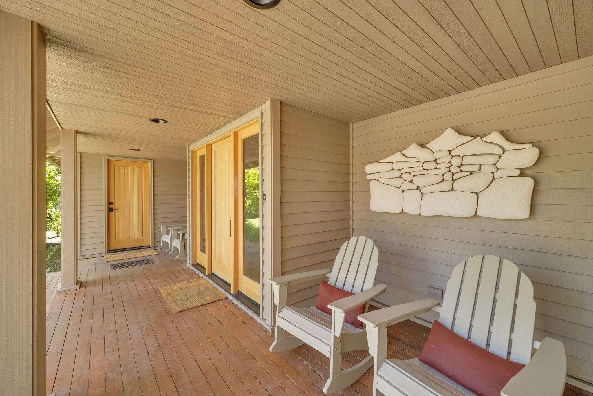 Un porche couvert avec un plancher en bois, deux fauteuils Adirondack, une porte en bois et un élément décoratif fixé au mur.