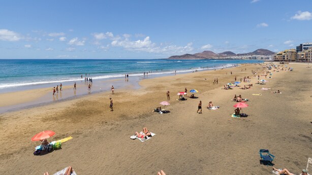 A wide sandy beach with people sunbathing and walking, a clear blue sky, and buildings in the background. - Charming Urban Getaway for Two (Las Palmas de Gran Canaria)
