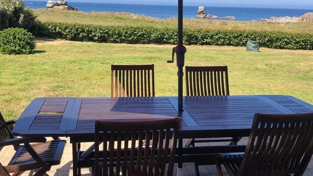 Una mesa y sillas de madera bajo un paraguas rojo, con vista a una zona verde y el océano con rocas.