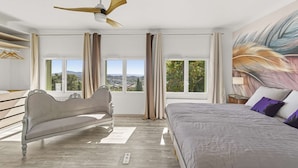 A bedroom with a bed, a ceiling fan, and a view of the city through large windows. - Panoramic sea view villa & heated pool (Toulon)