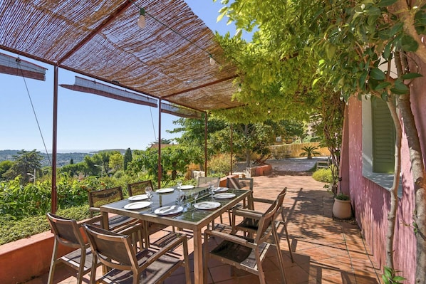 An outdoor dining area with a table set for four, surrounded by chairs, under a pergola with a view of the landscape.