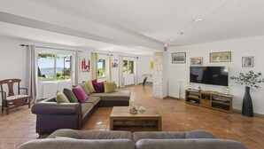 A spacious living room with a large sectional sofa, a coffee table, and a TV. There are large windows with curtains, a fireplace, and a stone accent wall. - Panoramic sea view villa & heated pool (Toulon)