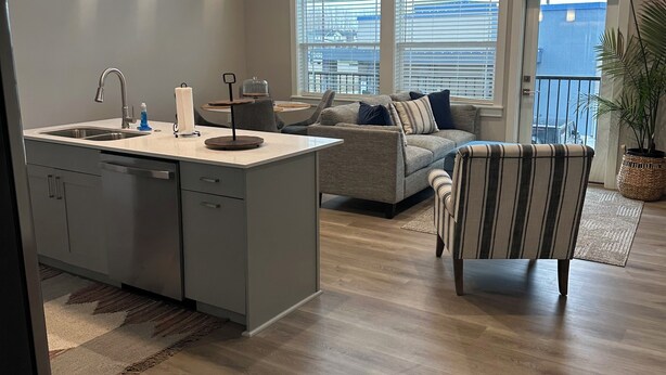 Une cuisine moderne avec un îlot central, un coin repas comprenant un canapé et un fauteuil, et de grandes fenêtres équipées de stores.