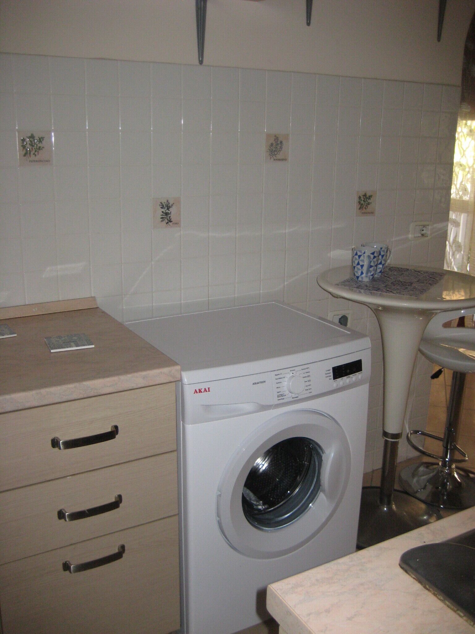 A compact kitchen with a front-loading washing machine, a small table with a cup, and a cabinet with drawers.