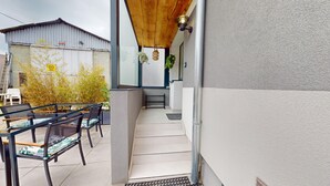 A narrow outdoor corridor with a metal staircase leading to a higher level. There are metal chairs and a table on the left, and a wall-mounted light fixture on the right.