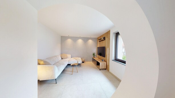 A modern living room with a sectional sofa, a TV mounted on a wooden panel, and a large window providing natural light.