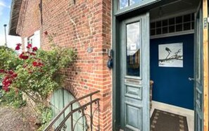 Un bâtiment en briques avec une porte bleue, un petit porche et un jardin où poussent des fleurs rouges.