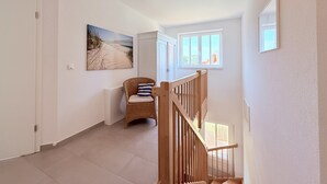A hallway with a wooden staircase, a wicker chair with a striped pillow, a beach-themed painting, and a window with a view of the outdoors. - Spacious, bright vacation home within walking distance of only 500m to the beach, with beautiful garden (Dassow)