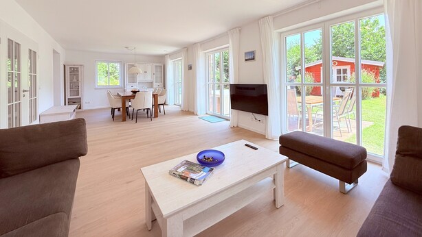 A spacious living room with a large TV, a dining area, and a view of a red house in the background. - Spacious, bright vacation home within walking distance of only 500m to the beach, with beautiful garden (Dassow)