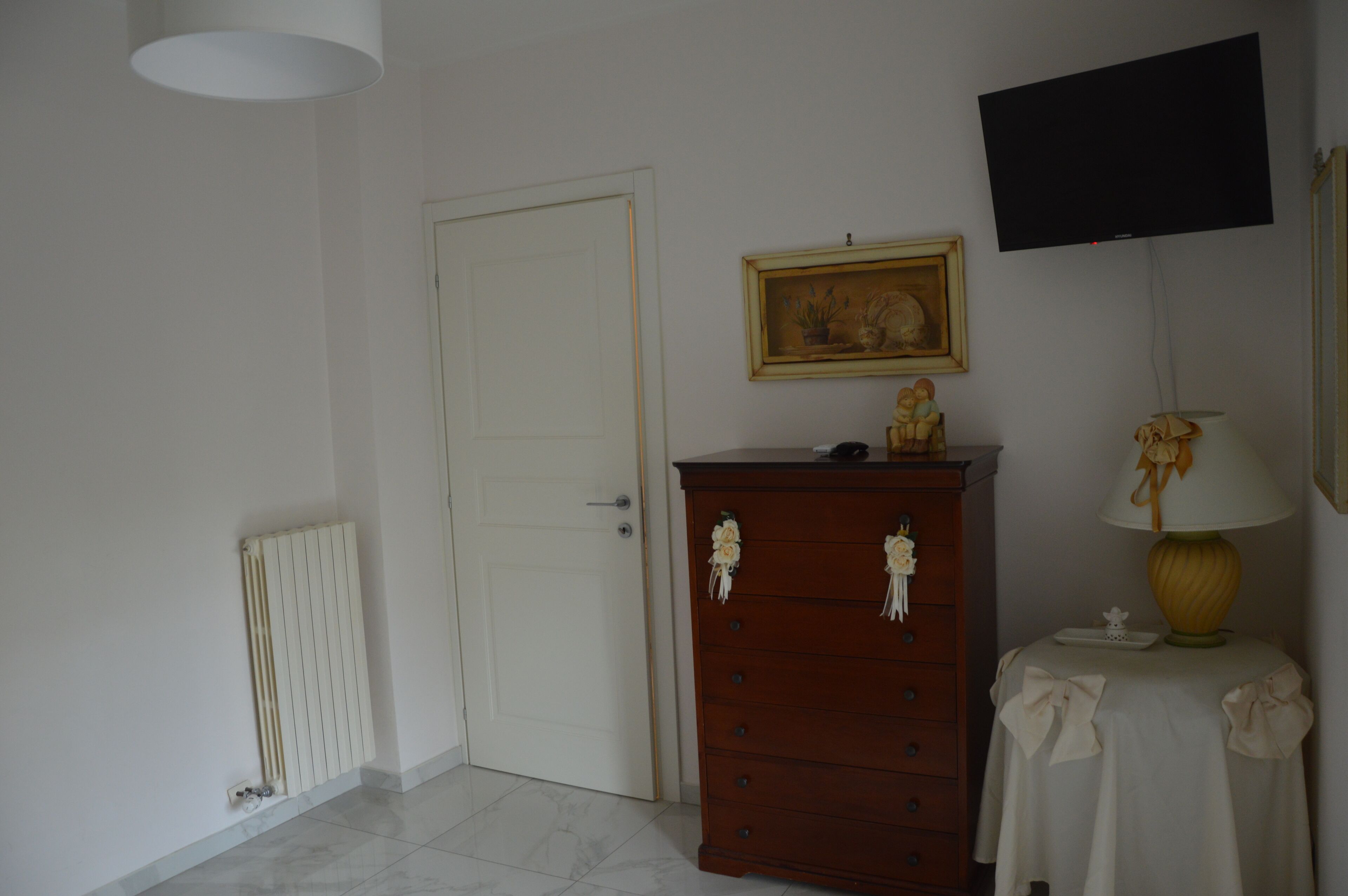 A room with a white door, a wooden dresser with decorative flowers, a framed picture on the wall, a table with a lamp, and a television mounted above.