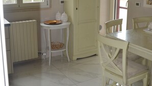 Ein Essbereich mit Tisch und Stühlen, ein kleiner runder Tisch mit einer Schüssel, ein hoher Schrank und ein Fenster mit Jalousien.