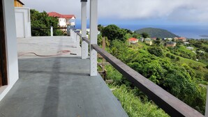 Un balcon doté d’un sol en béton et de rampes en bois, offrant une vue sur un paysage verdoyant et luxuriant, avec des maisons et une vue sur la mer.
