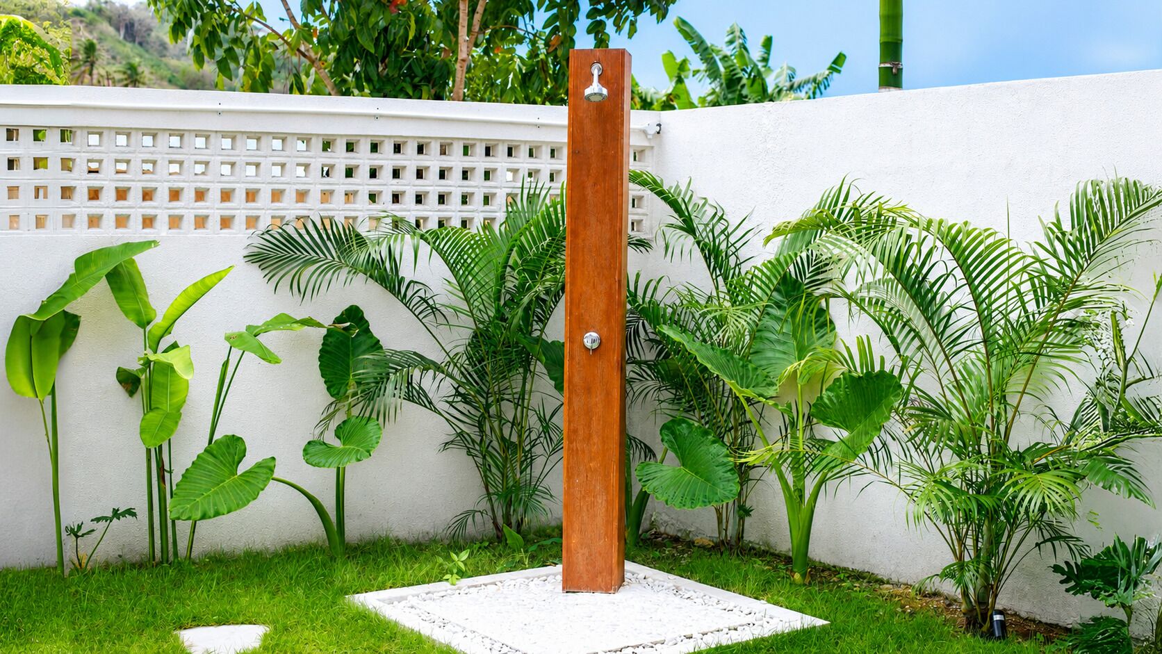 Un poste de madera con un cabezal de ducha, rodeado de plantas verdes exuberantes y una pared blanca.