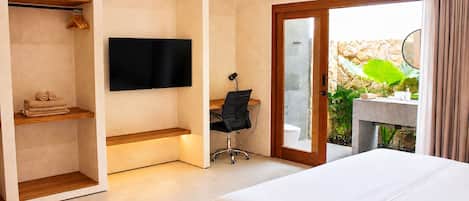 A hotel room with a bed, a desk with a chair, a television, a wardrobe, and a bathroom visible through a glass door.