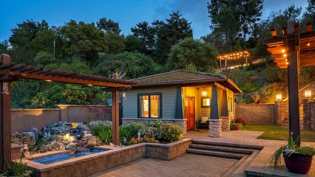 Une maison dotée d’une terrasse en pierre, d’une petite piscine et d’une pergola illuminée de guirlandes lumineuses.