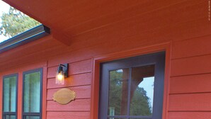 Una casa de madera roja con una puerta gris oscura, un letrero de bienvenida y una ventana con una lámpara.
