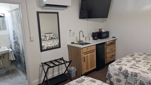Una habitación compacta con cama, televisión montada en la pared, kitchenette pequeño con lavabo y microondas, y ventana con vista a árboles.