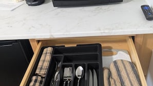 Una cocina con microondas, cafetera y un control remoto sobre la encimera. Debajo, un cajón contiene un juego de utensilios y una servilleta.