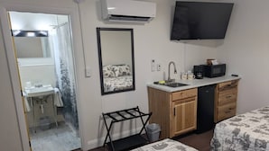 Un dormitorio pequeño con cama, una kitchenette, televisión y baño visible a través de una puerta abierta.