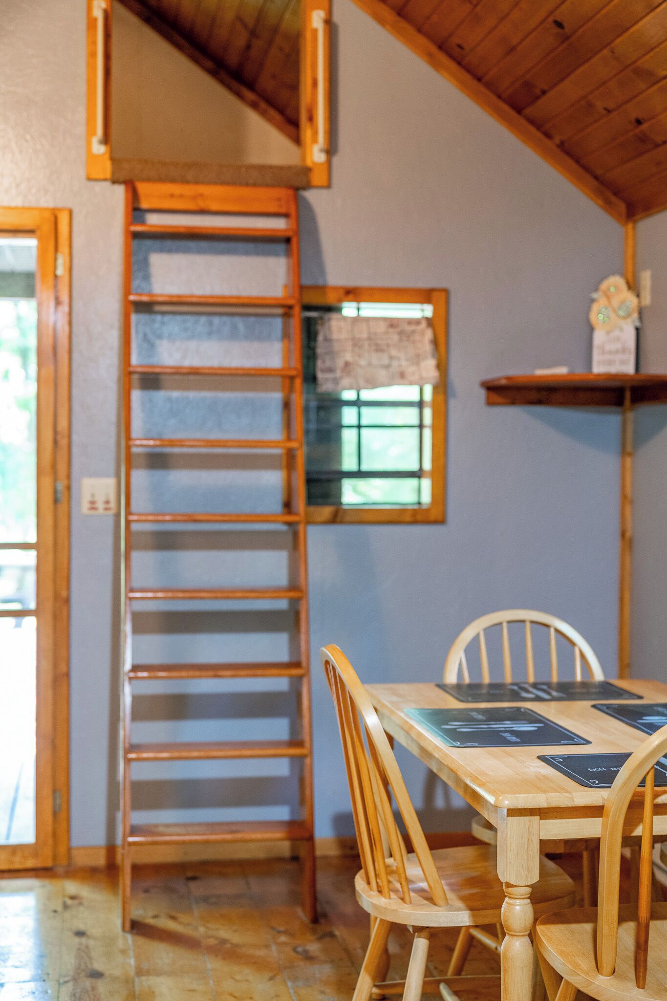 A wooden dining table with chairs, a wooden ladder, and a window with a curtain.
