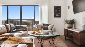 A modern living room with a leather sofa, a glass coffee table, and a view of the city through large windows. - The Crystal Palace 2, pet friendly 2 bedroom apartment (Jindabyne)
