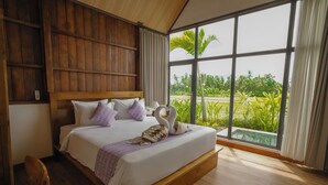 A spacious bedroom with a large bed, wooden headboard, and a view of lush greenery through large windows. - Kose Villa Ubud (Ubud)
