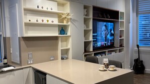 Moderni olohuone, jossa on televisio, koriste-esineillä täytetyt hyllyt, kitara ja keittiöalue, jossa on mikroaaltouuni.