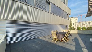 A rooftop terrace with a table and chairs, a closed roller shutter, and a view of a construction crane in the background.