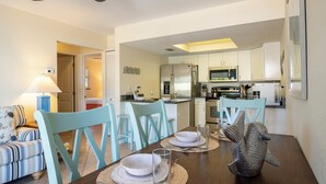 A dining area with a wooden table set for four, light blue chairs, and a fish decoration. A kitchen is visible in the background with white cabinets and stainless steel appliances. - Foxtail Palm: Steps from the beach (Holmes Beach)