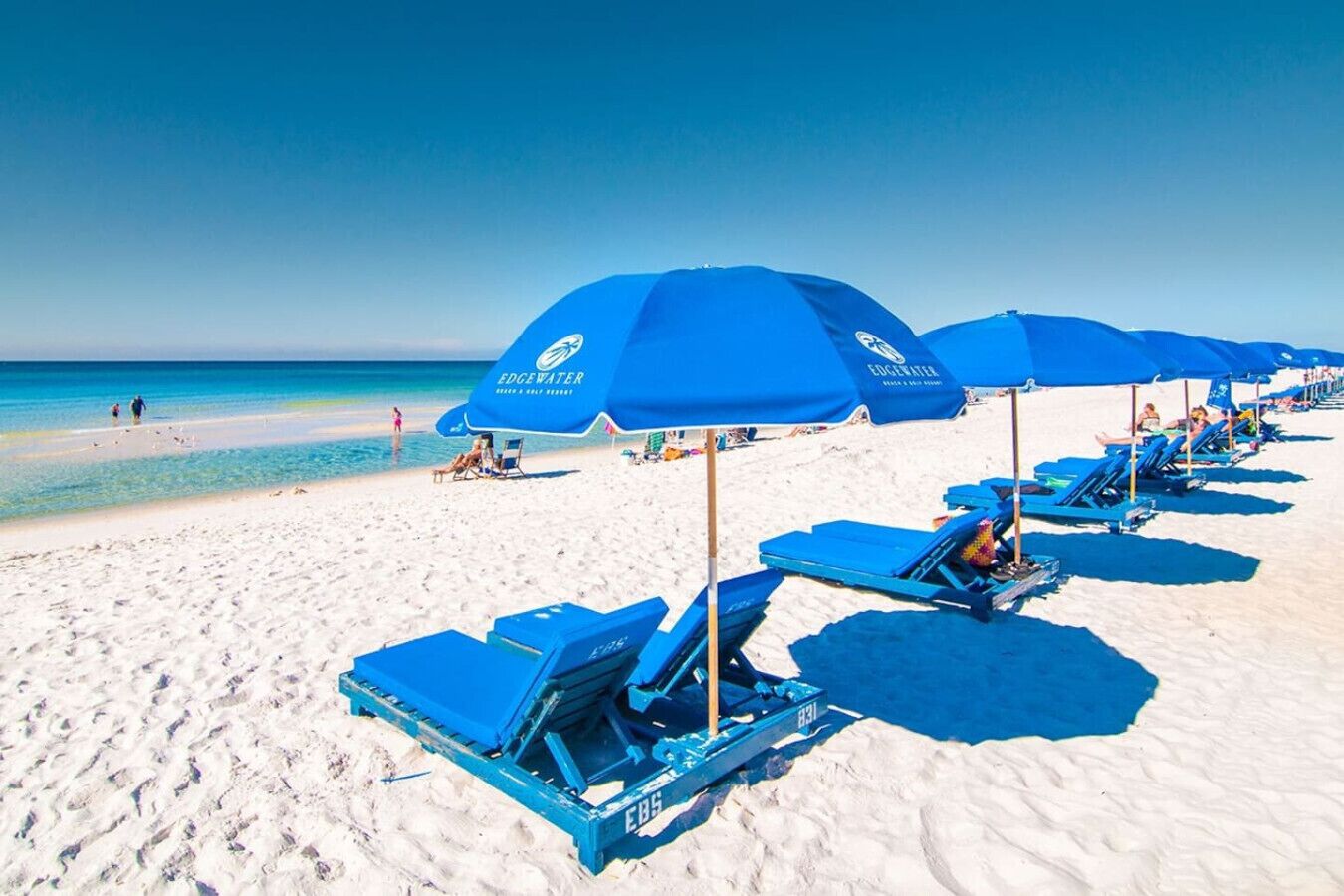 A beach with blue umbrellas and lounge chairs, a clear sky, and the ocean in the background.