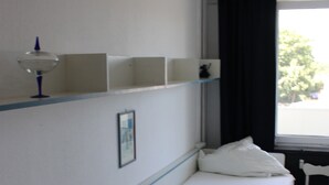 Ein Zimmer mit einem Einzelbett, einem Fenster, einem Sessel und einem Regal mit einer Lampe.