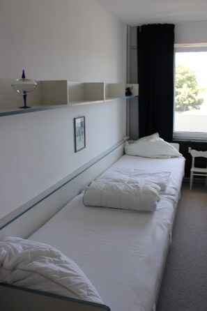 A single-bed room with a window, a chair, and a shelf with a lamp.