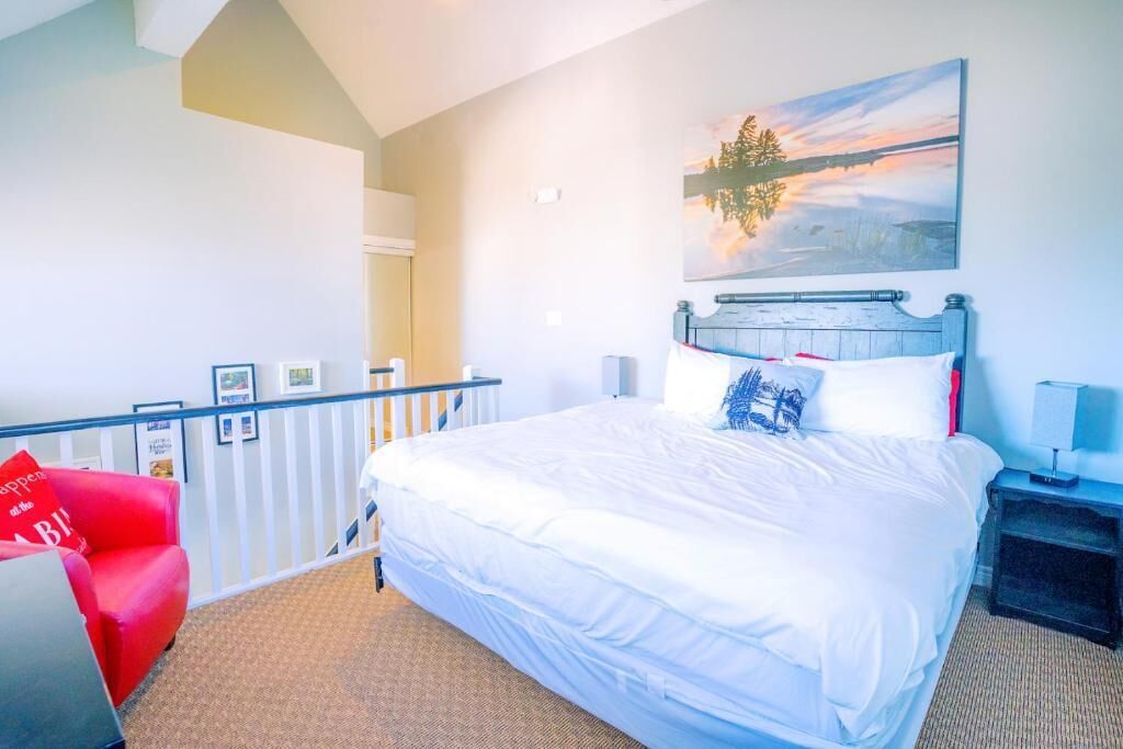 Quarto de hotel com uma cama grande, uma cadeira vermelha, um quadro de um pôr do sol e vista para o exterior.