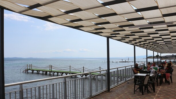 A covered outdoor seating area with a view of the sea, a dock, and a distant mountain range. - Address Küçükkuyu (Ayvacik)