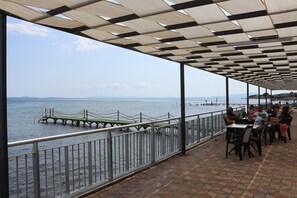 Et overdækket udendørs opholdsområde med udsigt til havet, en mole og fjerne bjerge.