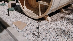 Une structure cylindrique en bois, dotée d’une porte et de bancs à l’intérieur, entourée d’arbres et de gravier.