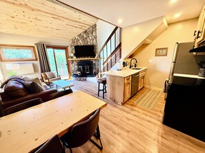 A modern interior with a dining area, kitchen, and living space, featuring a stone fireplace, wooden ceiling, and a staircase.