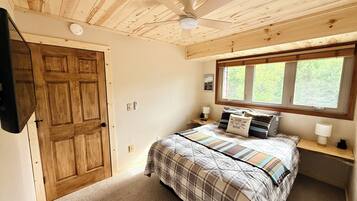 Een slaapkamer met een houten plafond, een bed met ruitdessin beddengoed, een raam met uitzicht en een deur.